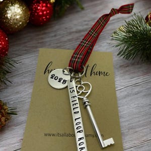 a custom new home ornament with a long silver tag with the street address hand stamped, a circle tag with the current year the home was purchased, a silver skeleton key, and a tartan plaid ribbon. set on a table with Christmas tree evergreen branches and soft twinkling lights and red ornaments
