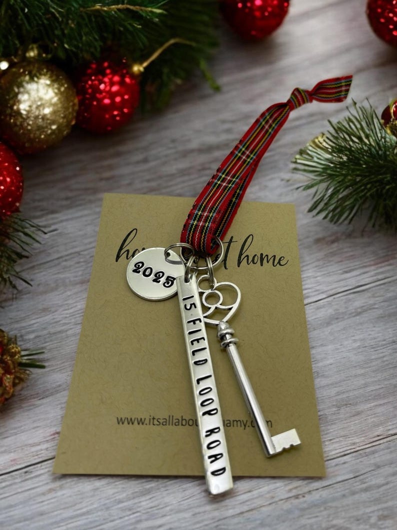 a custom new home ornament with a long silver tag with the street address hand stamped, a circle tag with the current year the home was purchased, a silver skeleton key, and a tartan plaid ribbon. set on a table with Christmas tree evergreen branches and soft twinkling lights and red ornaments