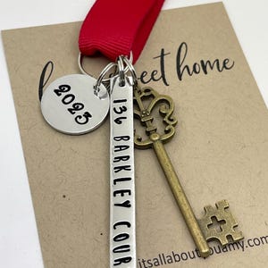 a custom new home ornament with a long silver tag with the street address hand stamped, a circle tag with the current year the home was purchased, a antique bronze skeleton key, and a red ribbon. set on a white background