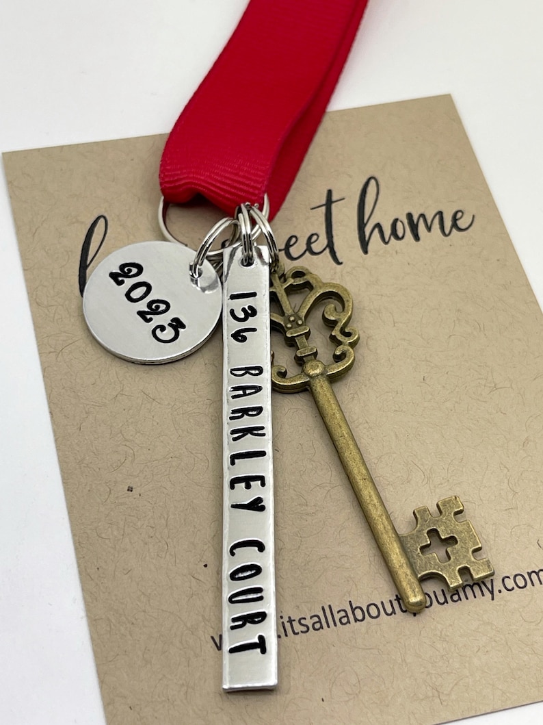 a custom new home ornament with a long silver tag with the street address hand stamped, a circle tag with the current year the home was purchased, a antique bronze skeleton key, and a red ribbon. set on a white background