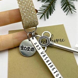 a custom new home ornament with a long silver tag with the street address hand stamped, a circle tag with the current year the home was purchased, a silver skeleton key, and a glitter ribbon. set on a white background with Christmas tree evergreen branches