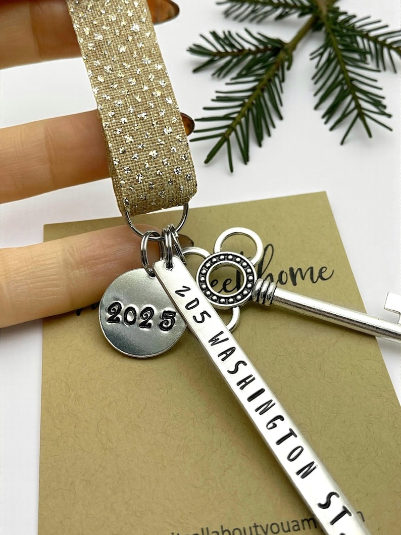 a custom new home ornament with a long silver tag with the street address hand stamped, a circle tag with the current year the home was purchased, a silver skeleton key, and a glitter ribbon. set on a white background with Christmas tree evergreen branches