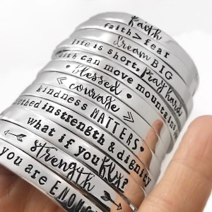 May include: A stack of silver metal bracelets with inspirational quotes engraved on them. The quotes include "faith > fear", "dream big", "life is short, pray hard", "blessed", "courage", "kindness matters", "strength & dignity", "what if you", "strength", and "you are enough".