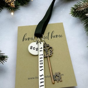 a custom new home ornament with a long silver tag with the street address hand stamped, a circle tag with the current year the home was purchased, a silver skeleton key, and a black satin ribbon. set on a white background with Christmas tree evergreen branches