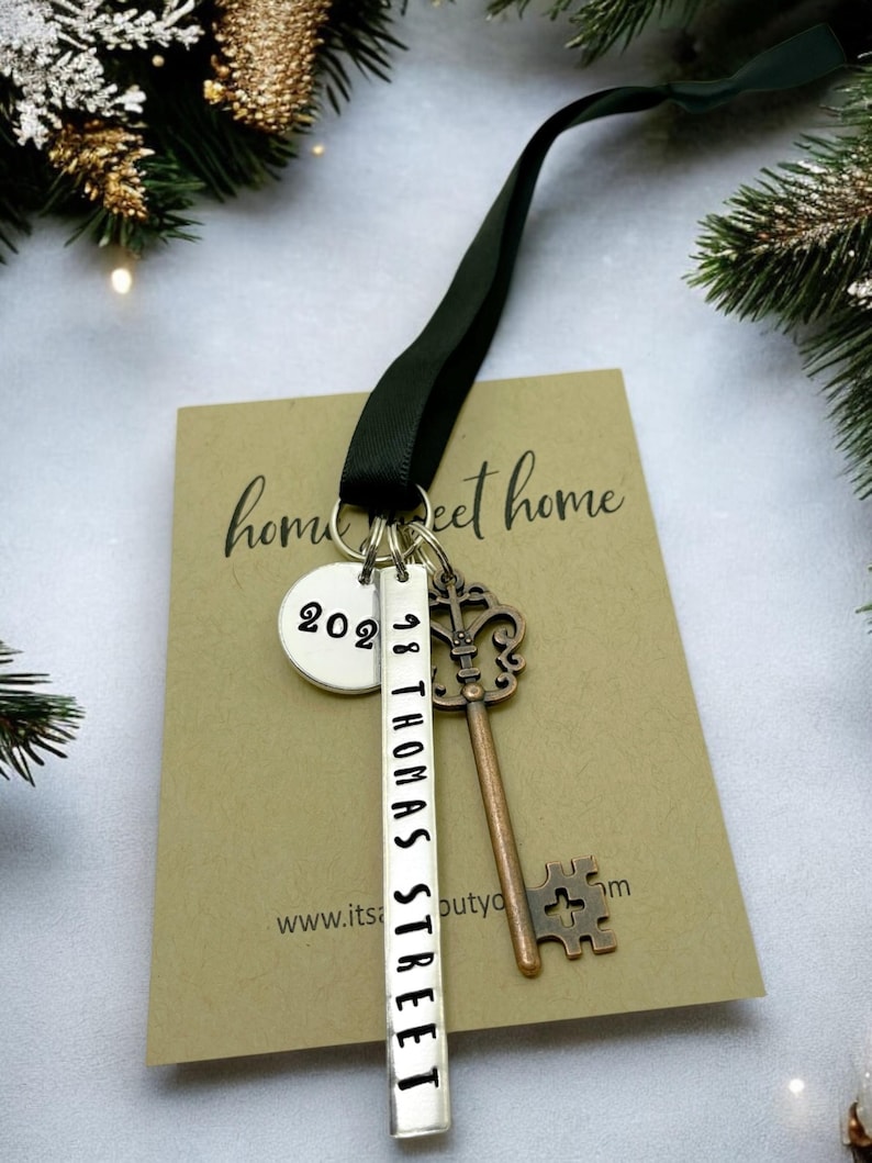 a custom new home ornament with a long silver tag with the street address hand stamped, a circle tag with the current year the home was purchased, a silver skeleton key, and a black satin ribbon. set on a white background with Christmas tree evergreen branches
