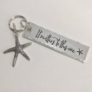 May include: A silver-toned metal keychain featuring a rectangular tag with the stamped text It matters to this one and a small starfish charm attached to a circular split ring. The rectangular tag has a textured, hammered edge finish.