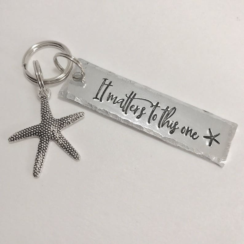 May include: A silver-toned metal keychain featuring a rectangular tag with the stamped text It matters to this one and a small starfish charm attached to a circular split ring. The rectangular tag has a textured, hammered edge finish.