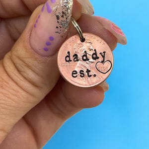 May include: A copper penny keychain stamped with "daddy est." and a heart symbol. The penny is attached to a silver keyring. The background is a solid light blue.