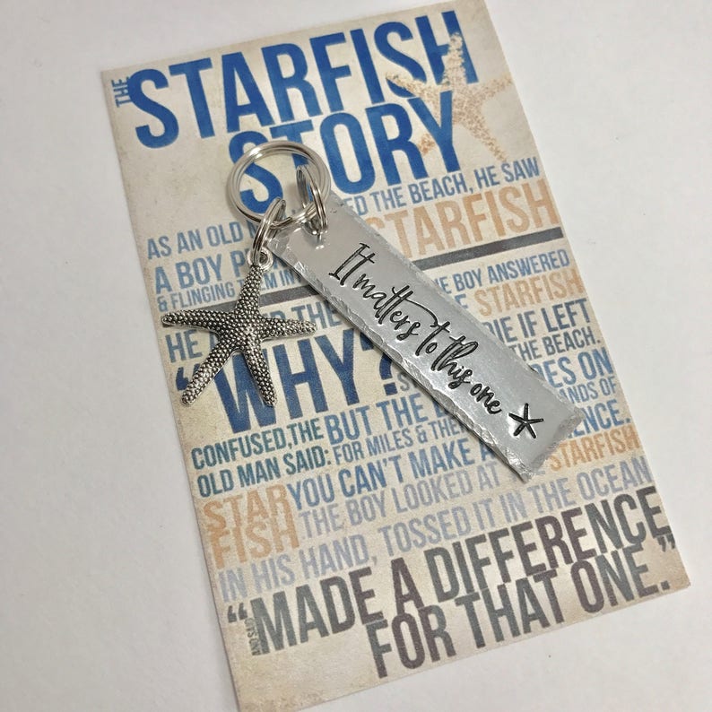May include: A silver-toned metal keychain featuring a rectangular tag stamped with the text It matters to this one and a small starfish charm. The keychain is attached to a rectangular card printed with the text The Starfish Story in blue and tan.