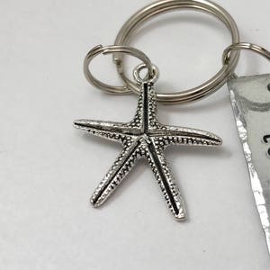 May include: A silver-toned metal starfish charm attached to a circular split key ring. The starfish features a textured, bumpy surface with a central line detail. A portion of a rectangular metal tag with partial black text is visible on the right.