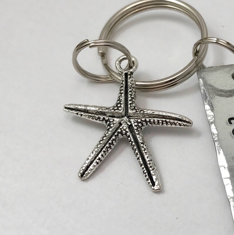 May include: A silver-toned metal starfish charm attached to a circular split key ring. The starfish features a textured, bumpy surface with a central line detail. A portion of a rectangular metal tag with partial black text is visible on the right.