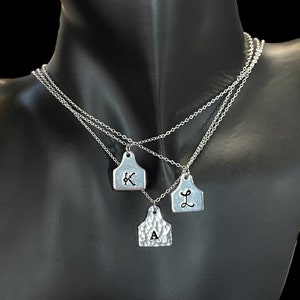 May include: Three silver-toned necklaces with pendant charms. Each charm is shaped like a tag, with the letters "K", "L", and "A" stamped on them. The necklaces are displayed on a black mannequin.