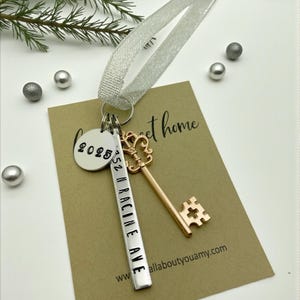 a custom new home ornament with a long silver tag with the street address hand stamped, a circle tag with the current year the home was purchased, a silver skeleton key, and a silver sparkle ribbon. set on a white background with Christmas tree evergreen branches