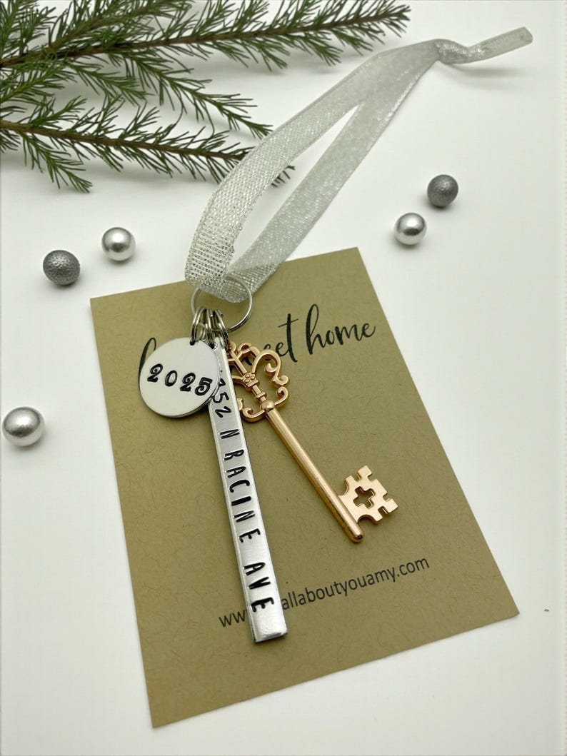 a custom new home ornament with a long silver tag with the street address hand stamped, a circle tag with the current year the home was purchased, a silver skeleton key, and a silver sparkle ribbon. set on a white background with Christmas tree evergreen branches