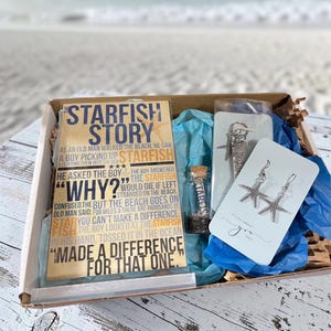 May include: A gift box with a beach theme. It contains a book titled "The Starfish Story", a small glass bottle, a starfish keychain, and a pair of starfish earrings. The box is lined with blue tissue paper.