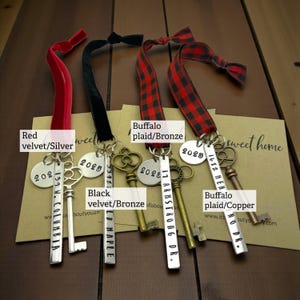 4 custom new home ornaments with a long silver tag with the street address hand stamped, a circle tag with the current year the home was purchased, a silver skeleton key, and a plush red velvet ribbon. set on dark wood background, shown with red velvet, black velvet, and buffalo plaid ribbons