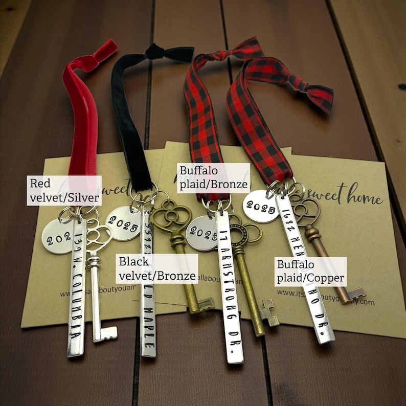4 custom new home ornaments with a long silver tag with the street address hand stamped, a circle tag with the current year the home was purchased, a silver skeleton key, and a plush red velvet ribbon. set on dark wood background, shown with red velvet, black velvet, and buffalo plaid ribbons