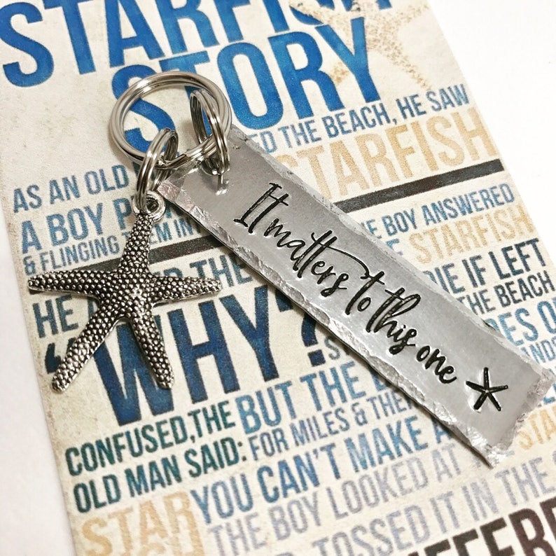 May include: A silver-toned metal keychain featuring a rectangular tag stamped with the text "It matters to this one" and a small starfish charm. The keychain is attached to a circular metal ring, resting against a card printed with text.