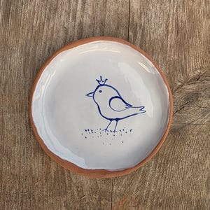 Pode incluir: Um prato redondo de cerâmica com uma borda cor de terracota e um centro esmaltado em branco. O interior apresenta uma ilustração a tinta azul desenhada à mão de um pequeno pássaro com uma coroa, sobre uma linha pontilhada na base.