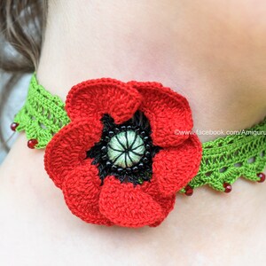 Puede incluir: Una flor de amapola roja de ganchillo con un centro negro est&aacute; unida a un collar de ganchillo verde con cuentas rojas. El collar se lleva alrededor del cuello de una persona.