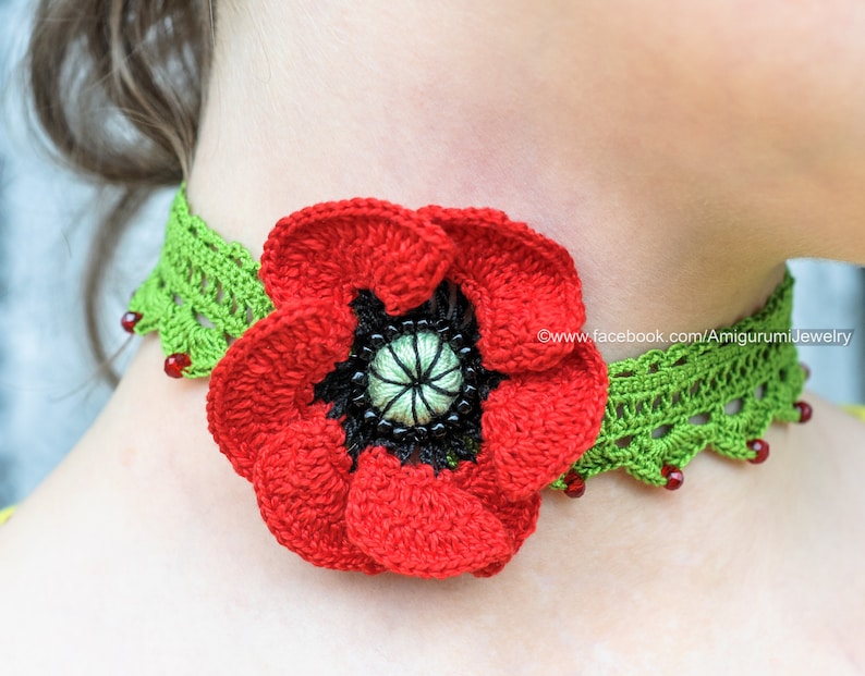 Puede incluir: Una flor de amapola roja de ganchillo con un centro negro est&aacute; unida a un collar de ganchillo verde con cuentas rojas. El collar se lleva alrededor del cuello de una persona.