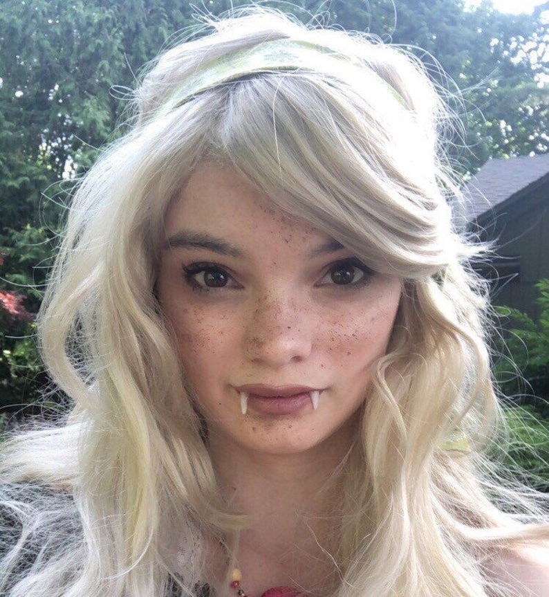 May include: A woman with long, blonde hair wearing a green headband and a necklace. She is wearing vampire fangs and has freckles on her face.