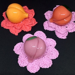 May include: Three glass peach-shaped ornaments, each sitting on a crocheted flower-shaped coaster. The coasters are pink, orange, and light pink.