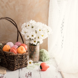 A basket of peach-shaped glass smoking pipes sits next to a vase of flowers near window. The image gives cottage core aesthetics and the peaches are a variety of peachy and pink colors.