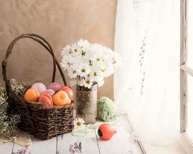 A basket of peach-shaped glass smoking pipes sits next to a vase of flowers near window. The image gives cottage core aesthetics and the peaches are a variety of peachy and pink colors.