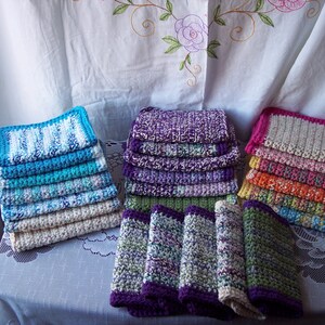 May include: A stack of colorful crocheted dishcloths in various patterns and colors, including blue, purple, pink, orange, and green. The dishcloths are stacked on a white tablecloth.