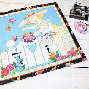 Könnte beinhalten: Ein farbenfrohes Stoffpanel mit einer skurrilen Illustration eines Mädchens, das Blumen gießt. Das Mädchen trägt ein gelbes Kleid mit Blumenmuster und einen rosa Hut. Das Panel zeigt einen weißen Lattenzaun mit einer herzförmigen Öffnung und einem blauen Himmel als Hintergrund. Das Panel ist mit einem schwarz-weißen Karomuster eingefasst.