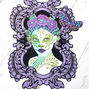 Może przedstawiać: Ozdobna fioletowa rama z czarnym tłem przedstawia kolorową ilustrację kobiety z makijażem czaszki cukrowej. Ma na sobie wianek kwiatowy, a na ramie siedzi motyl.