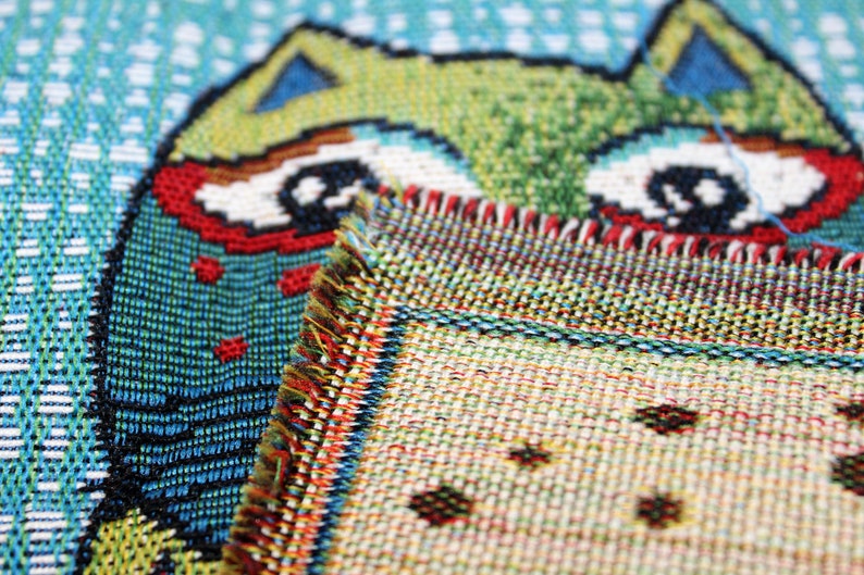 May include: Close-up of a colorful woven tapestry featuring a cartoon owl with large eyes. The owl is partially obscured by a second piece of tapestry with a geometric pattern.