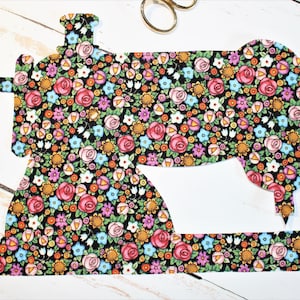 Könnte beinhalten: Ein aus Stoff ausgeschnittener Blumenmuster in Form einer Nähmaschine. Der Stoff hat einen schwarzen Hintergrund mit einem sich wiederholenden Muster aus rosa, roten, gelben, blauen und grünen Blumen.