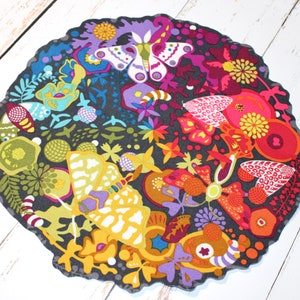 May include: A round, decorative mat with a dark gray background and a colorful floral and butterfly design. The design includes shades of purple, yellow, orange, red, and blue. The mat has a scalloped edge.