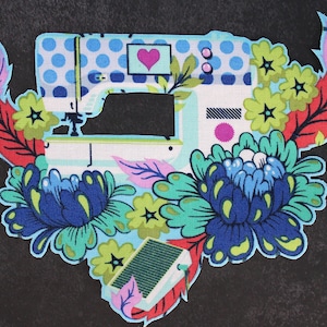 Puede incluir: Un parche colorido con un diseño de máquina de coser, un corazón y elementos florales azules, verdes y rojos. La máquina de coser tiene un patrón de lunares azules y una base blanca. El parche está sobre un fondo gris oscuro.