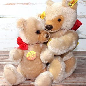May include: Two plush teddy bears in a warm beige color, one embracing the other. Each bear has a red ribbon tied around its neck and a yellow tag on its ear. The bears have brown eyes and noses, and are set against a light wood background.