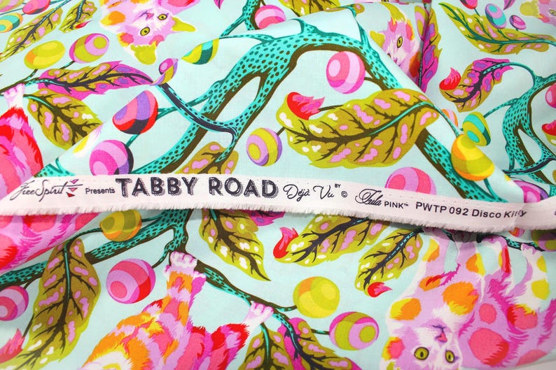 K&ouml;nnte beinhalten: Stoff mit einem farbenfrohen Blumen- und Katzenmuster. Der Stoff zeigt rosa, gelbe und lila Blumen sowie Katzen in verschiedenen Posen. Der Hintergrund ist hellblau. Der Text "TABBY ROAD" ist auf einem wei&szlig;en Band aufgedruckt.