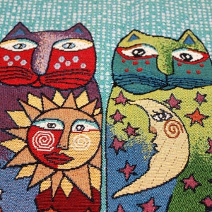 May include: A tapestry featuring two cats with whimsical faces, a sun with a smiling face, and a crescent moon with a face. The background is a blue and green pattern with stars.