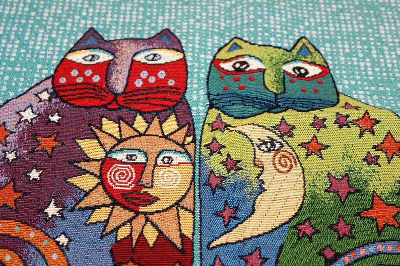 May include: A tapestry featuring two cats with whimsical faces, a sun with a smiling face, and a crescent moon with a face. The background is a blue and green pattern with stars.