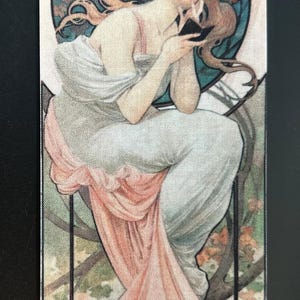 May include: A rectangular bookmark with an Art Nouveau design. It depicts a woman in a flowing white and pink gown, holding a small object. The background features a blue circle with floral and foliage elements.