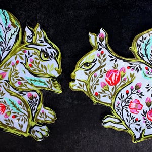 Może przedstawiać: Dwa dzieła sztuki w kształcie wiewiórki z motywami kwiatowymi. Wiewiórki są białe z czerwonymi, różowymi i zielonymi wzorami kwiatowymi i złotym konturem. Tło jest czarne.