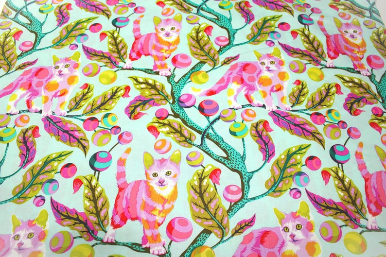 K&ouml;nnte beinhalten: Ein Stoff mit einem sich wiederholenden Muster aus rosa und orangefarbenen Katzen, die auf t&uuml;rkisfarbenen &Auml;sten mit bunten Beeren und Bl&auml;ttern sitzen. Die Katzen haben gelbe Ohren und sind vor einem hellblauen Hintergrund platziert.