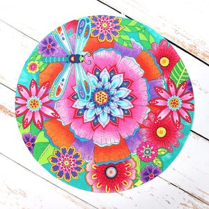 May include: A round, colorful floral design featuring a large pink flower at the center, surrounded by smaller flowers in shades of red, purple, and pink. A blue dragonfly is positioned over the central flower. The design is set against a turquoise background.