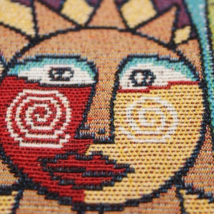 May include: Close-up of a woven tapestry featuring a stylized sun with a red, white, and yellow face. The sun has a spiral design on the left side of its face.