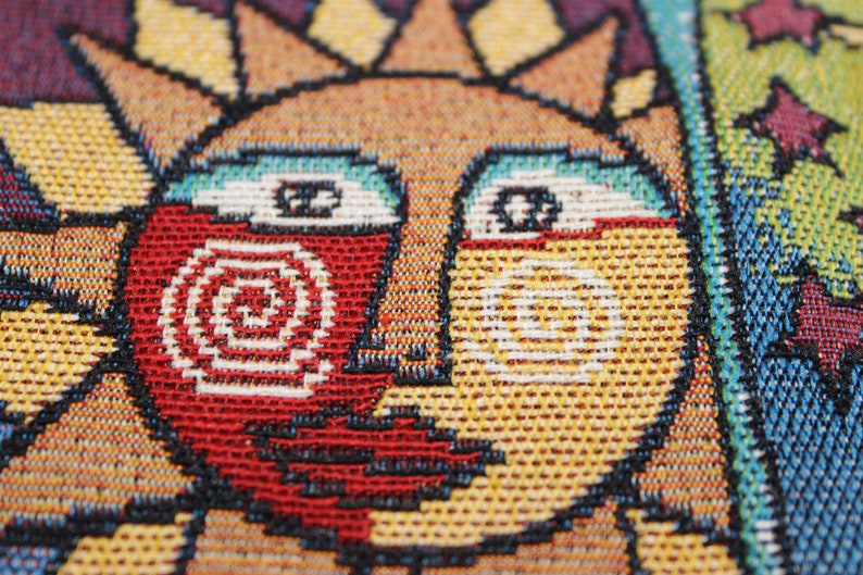 May include: Close-up of a woven tapestry featuring a stylized sun with a red, white, and yellow face. The sun has a spiral design on the left side of its face.