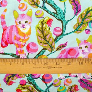 K&ouml;nnte beinhalten: Stoff mit einem skurrilen Design mit zwei stilisierten Katzen in rosa, orange und gelben Streifen und Flecken. Die Katzen sind vor einem hellblauen Hintergrund mit bunten, gemusterten Kugeln und Bl&auml;ttern platziert. Unten befindet sich ein Holzlineal.