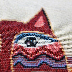 May include: Close-up of a red, white, and blue woven tapestry featuring a cat with large, black eyes.