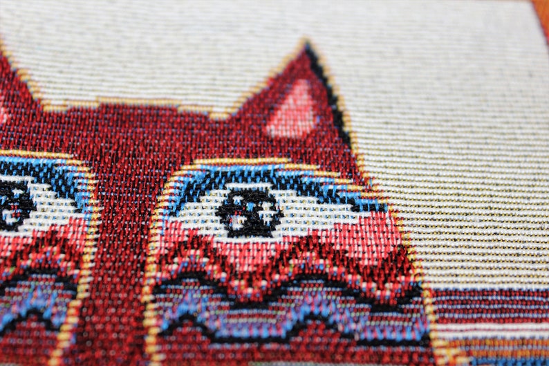 May include: Close-up of a red, white, and blue woven tapestry featuring a cat with large, black eyes.