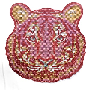May include: A decorative tiger head design in shades of red, pink, yellow, and white. The artwork features a mosaic-like pattern, with the tiger's face and features clearly defined. The eyes are light blue, and the overall design is symmetrical.